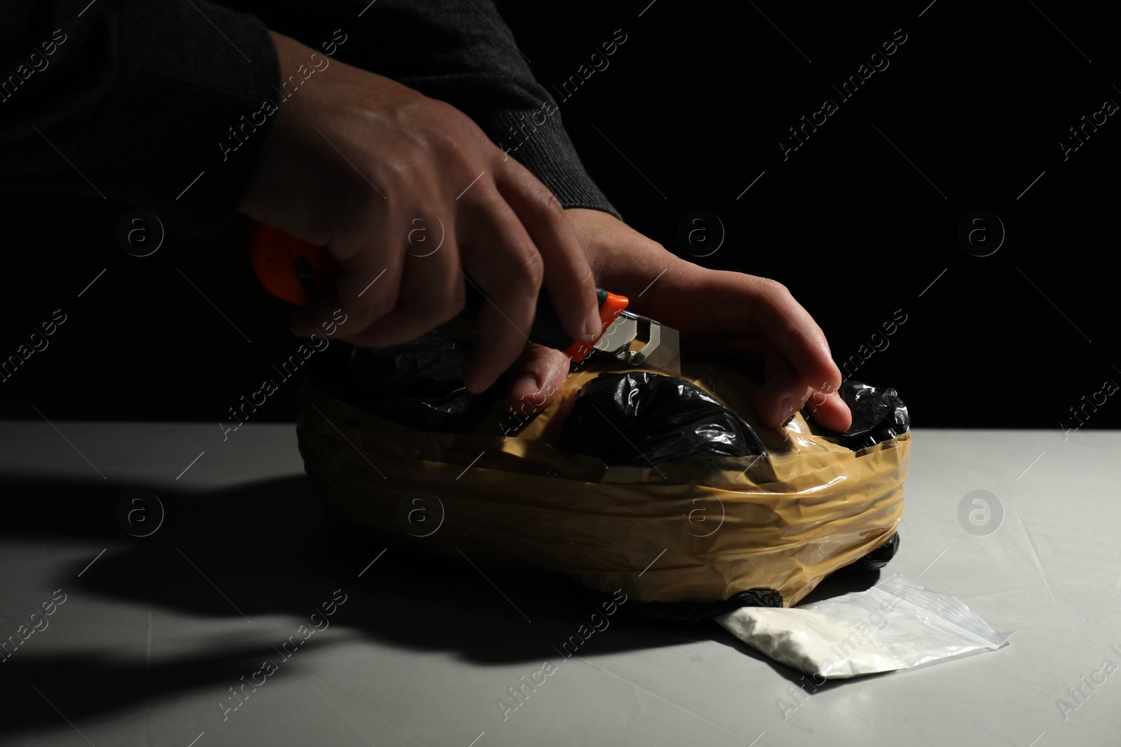 Photo of Smuggling and drug trafficking. Man opening package of narcotics with box cutter at light table against black background, closeup
