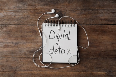 Notebook with words DIGITAL DETOX and earphones on wooden background, flat lay