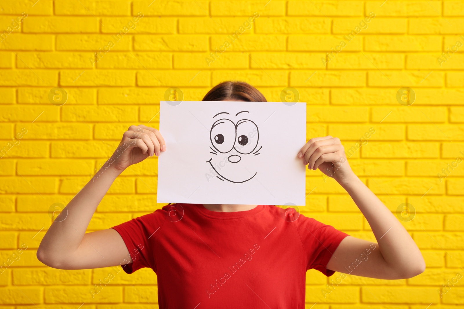 Photo of Woman hiding behind sheet of paper with happy face near yellow brick wall