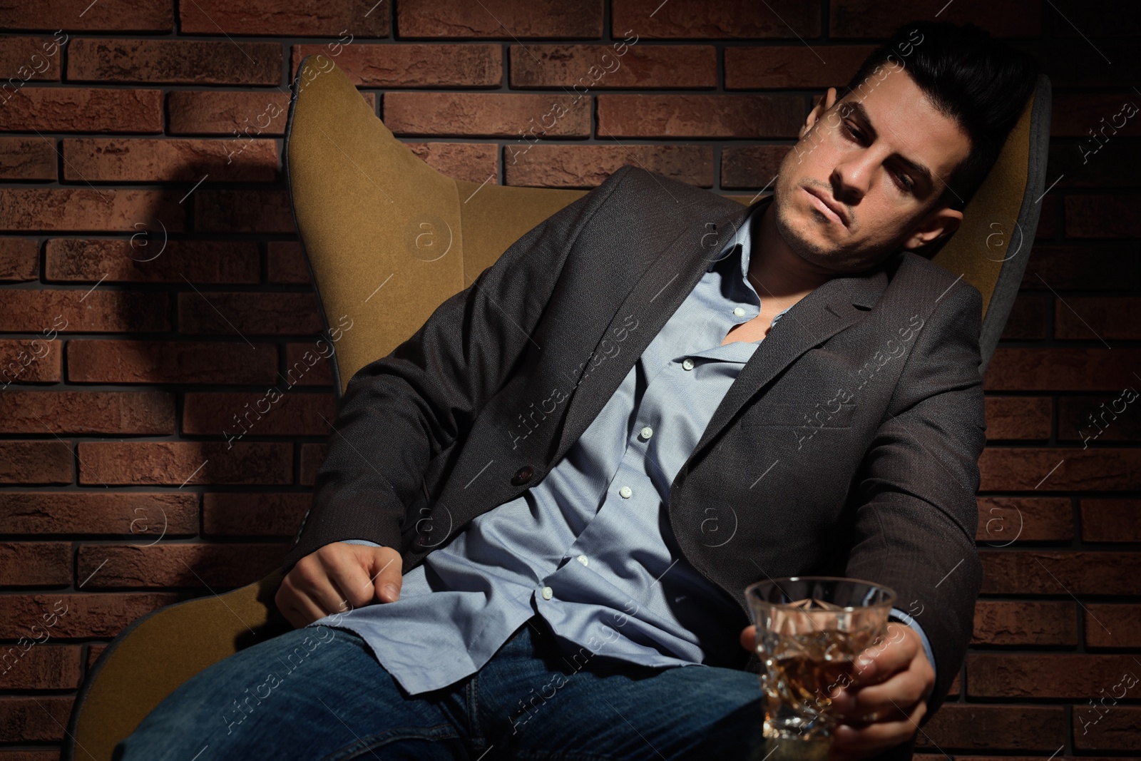 Photo of Addicted man with glass of alcoholic drink in armchair near red brick wall