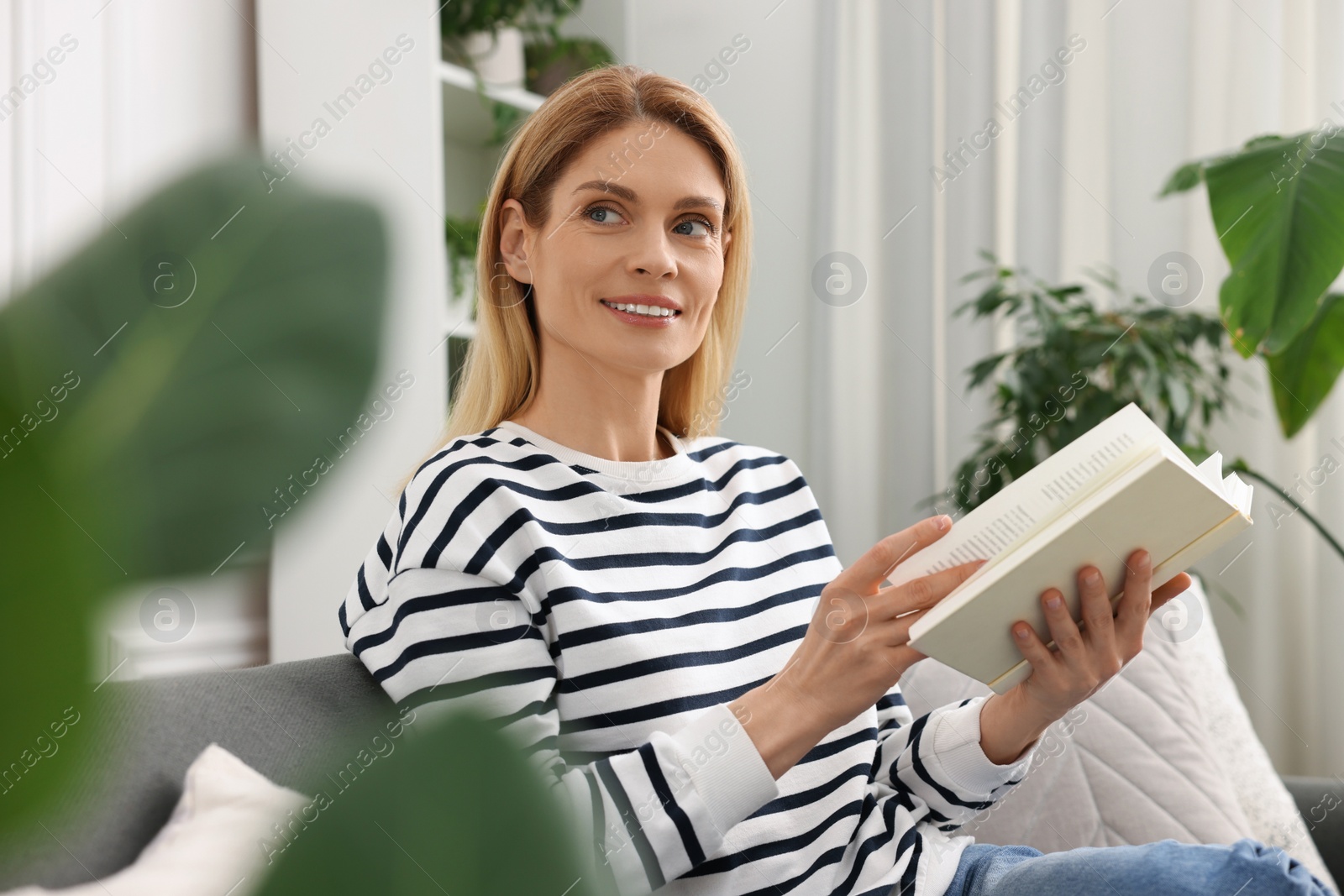 Photo of Woman reading book on sofa near beautiful potted houseplants at home