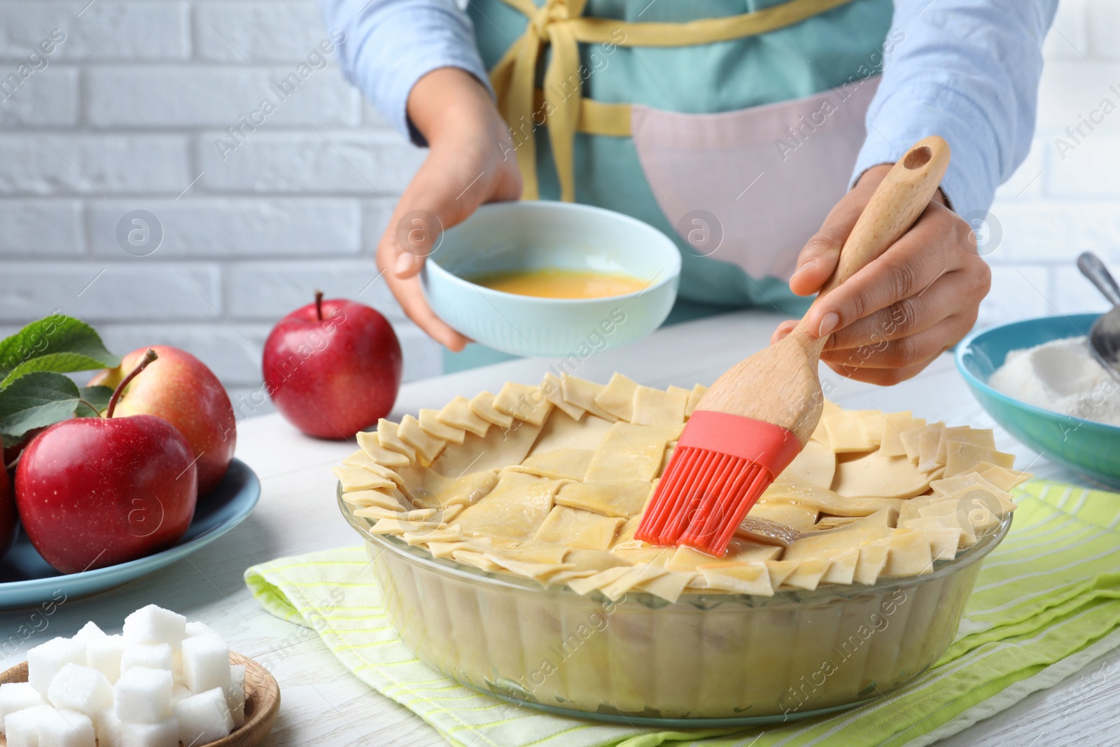 Photo of Woman spreading egg yolk onto raw apple pie at white wooden table, closeup