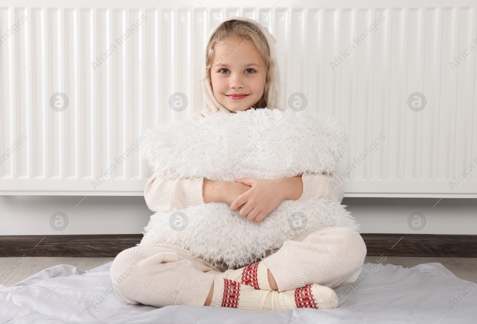 Photo of Little girl with pillow near heating radiator indoors