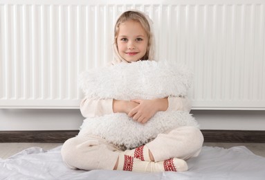 Photo of Little girl with pillow near heating radiator indoors