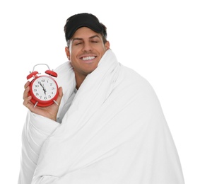 Man in sleeping mask wrapped with blanket holding alarm clock on white background