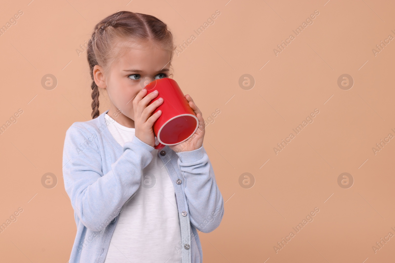 Photo of Cute girl drinking beverage from red ceramic mug on beige background, space for text