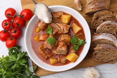 Delicious goulash in bowl, bread and ingredients on white textured table, flat lay