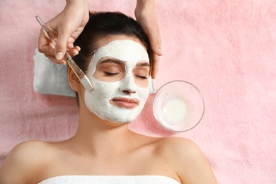 Cosmetologist applying white mask onto woman's face in spa salon, top view