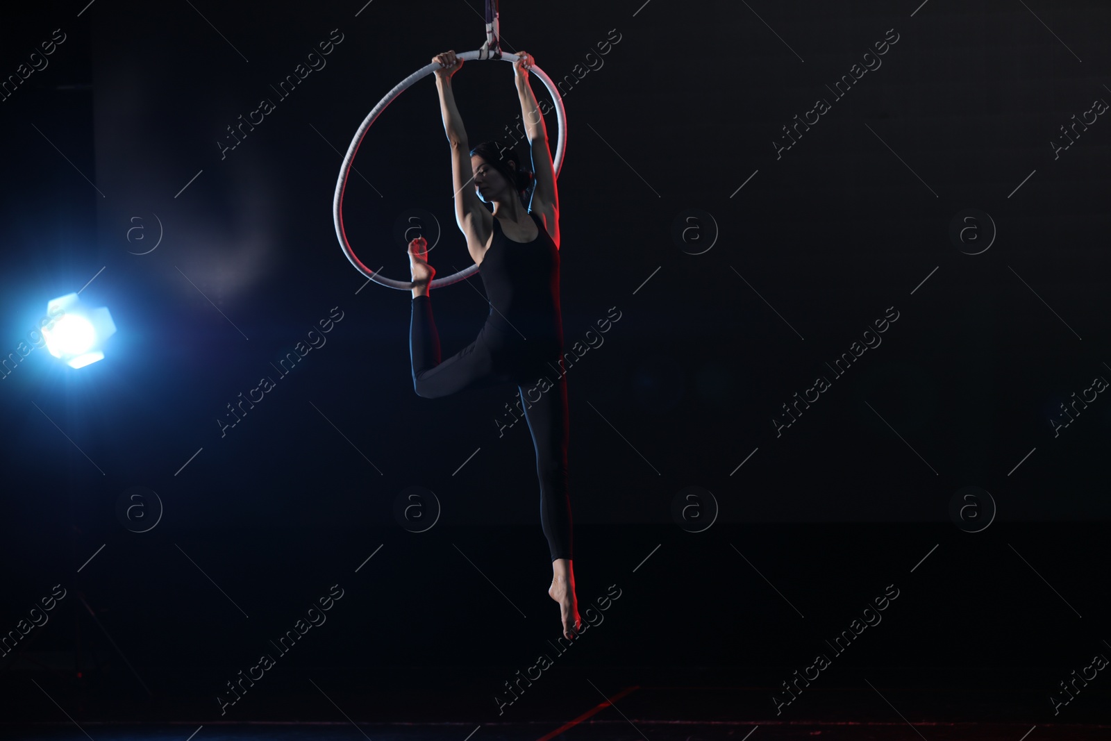 Photo of Young woman performing acrobatic element on aerial ring against dark background