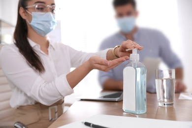 Photo of Office worker in protective mask using sanitizer at table, focus on hands. Personal hygiene during COVID-19 pandemic