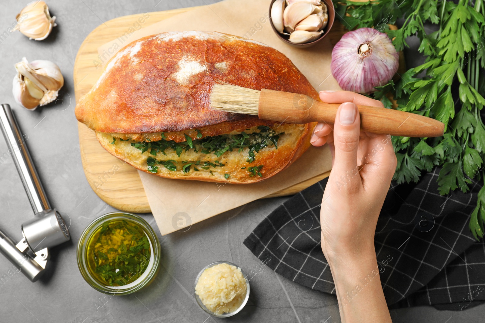 Photo of Woman brushing delicious homemade bread with herbs and garlic at grey table, top view