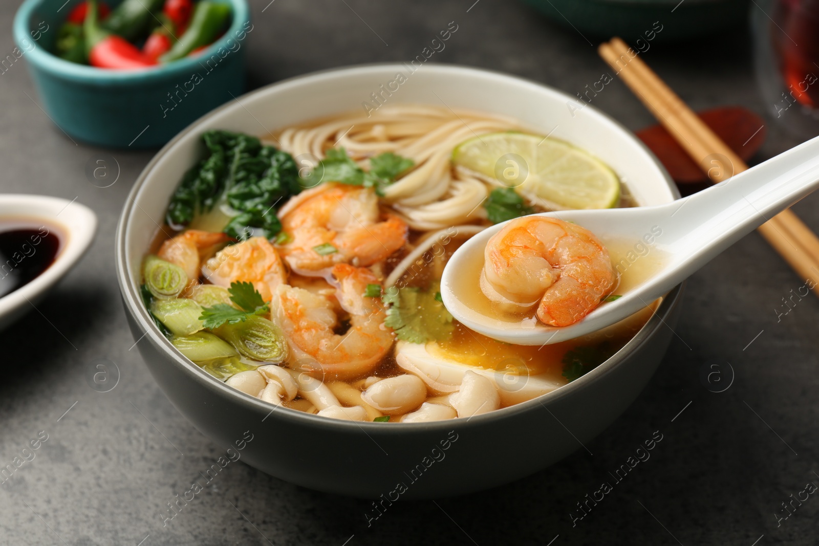 Photo of Eating delicious ramen with shrimps from bowl with spoon at grey table, closeup. Noodle soup