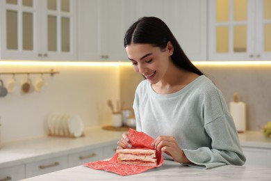 Photo of Happy woman packing sandwich into beeswax food wrap at table in kitchen