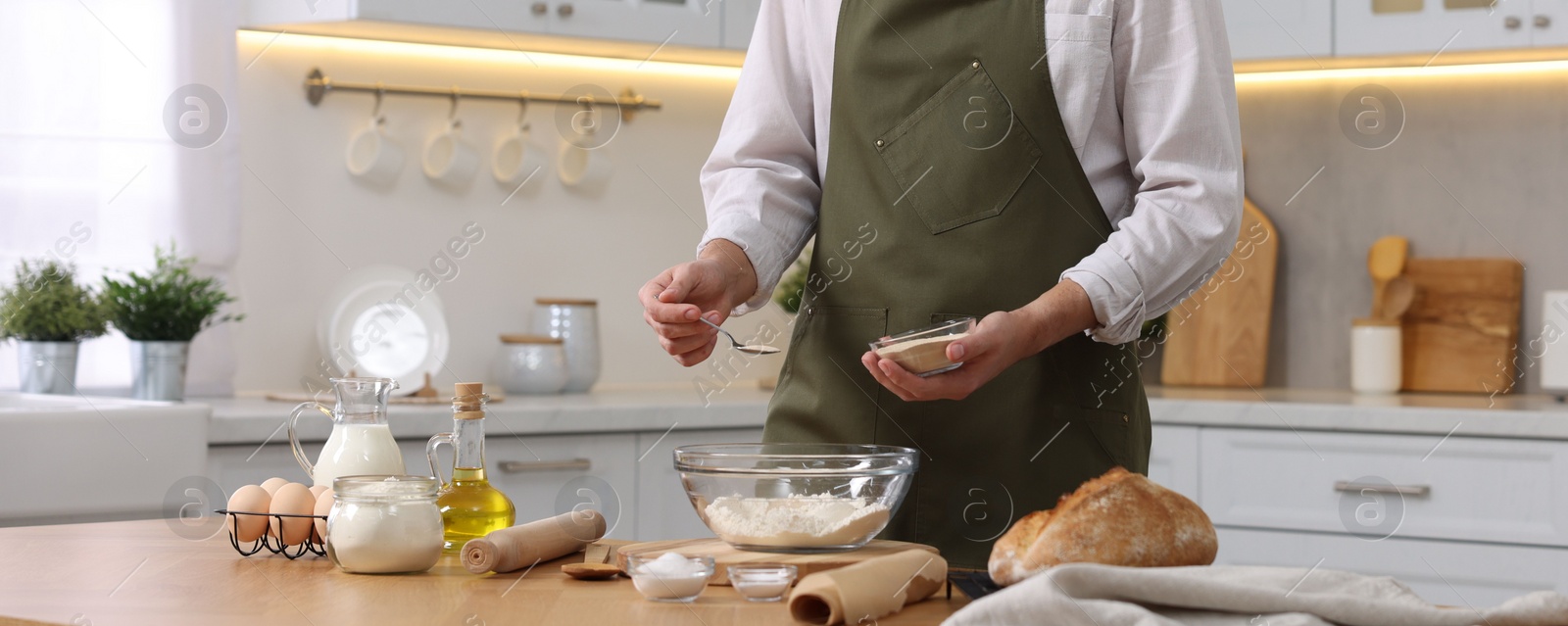 Photo of Making bread. Man putting dry yeast into bowl with flour at wooden table in kitchen, closeup