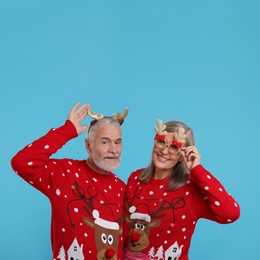 Photo of Senior couple in Christmas sweaters, reindeer headband and funny glasses on light blue background