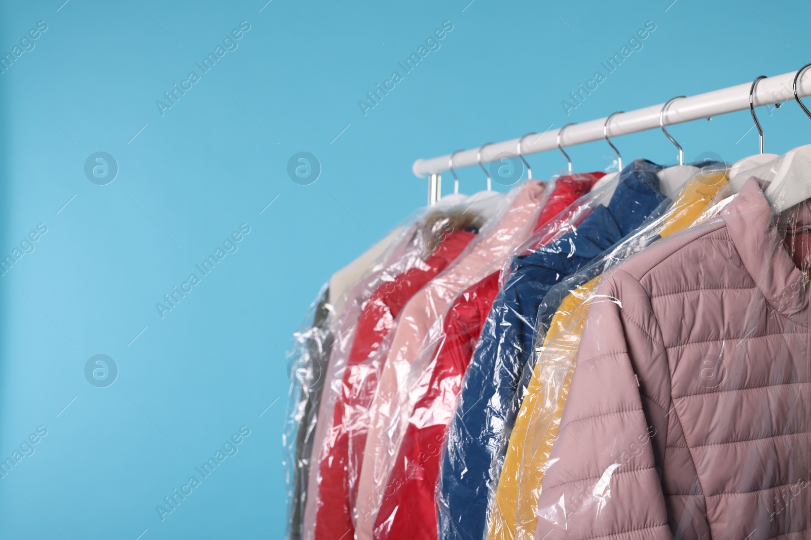 Photo of Dry-cleaning service. Many different clothes in plastic bags hanging on rack against light blue background, space for text