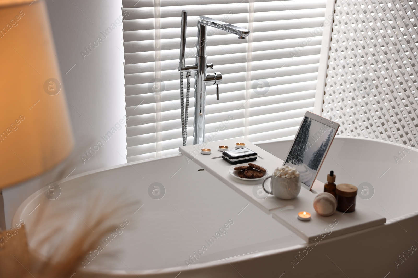 Photo of White wooden tray with tablet, spa products and burning candles on bathtub in bathroom