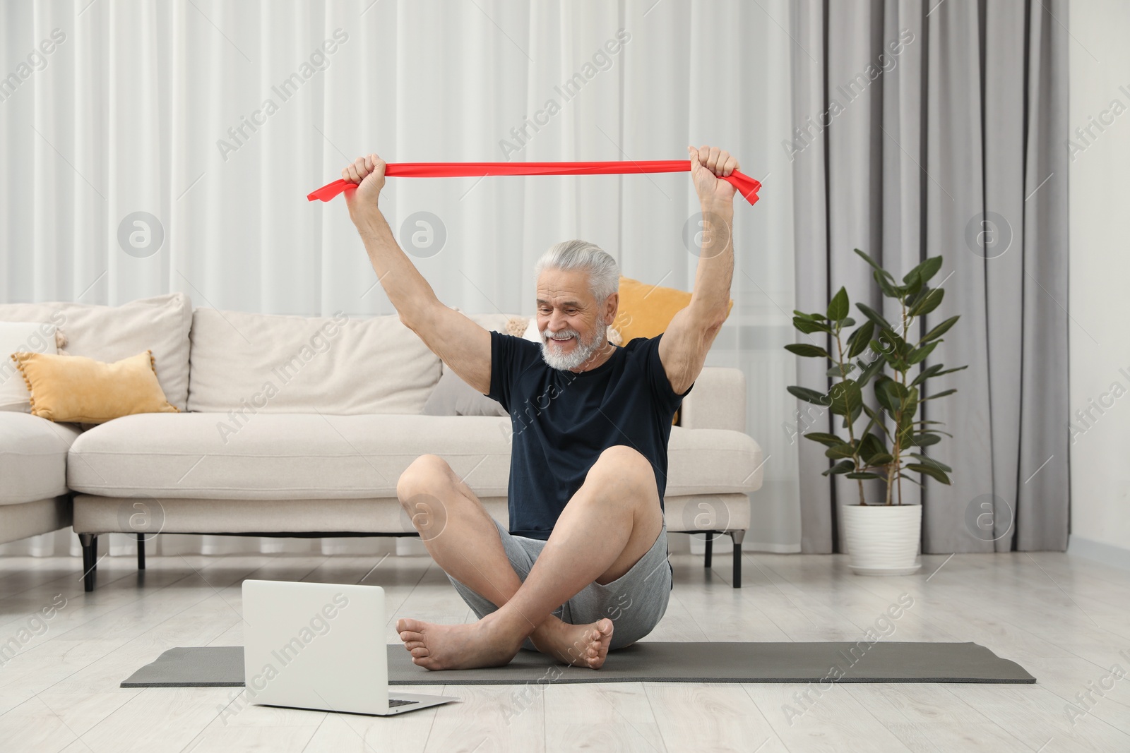 Photo of Senior man doing exercise with fitness elastic band near laptop on mat at home