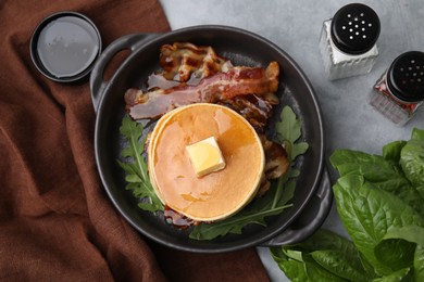 Tasty pancakes with butter, fried bacon and fresh arugula on grey table, flat lay