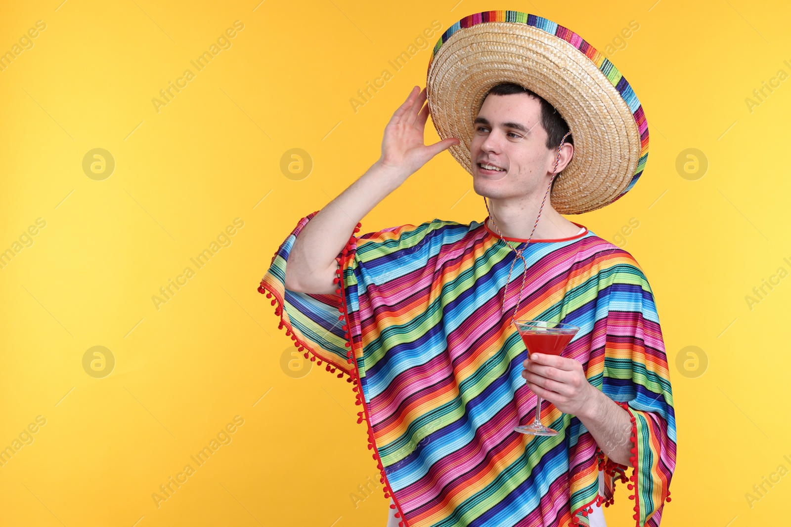 Photo of Young man in Mexican sombrero hat and poncho with cocktail on yellow background. Space for text