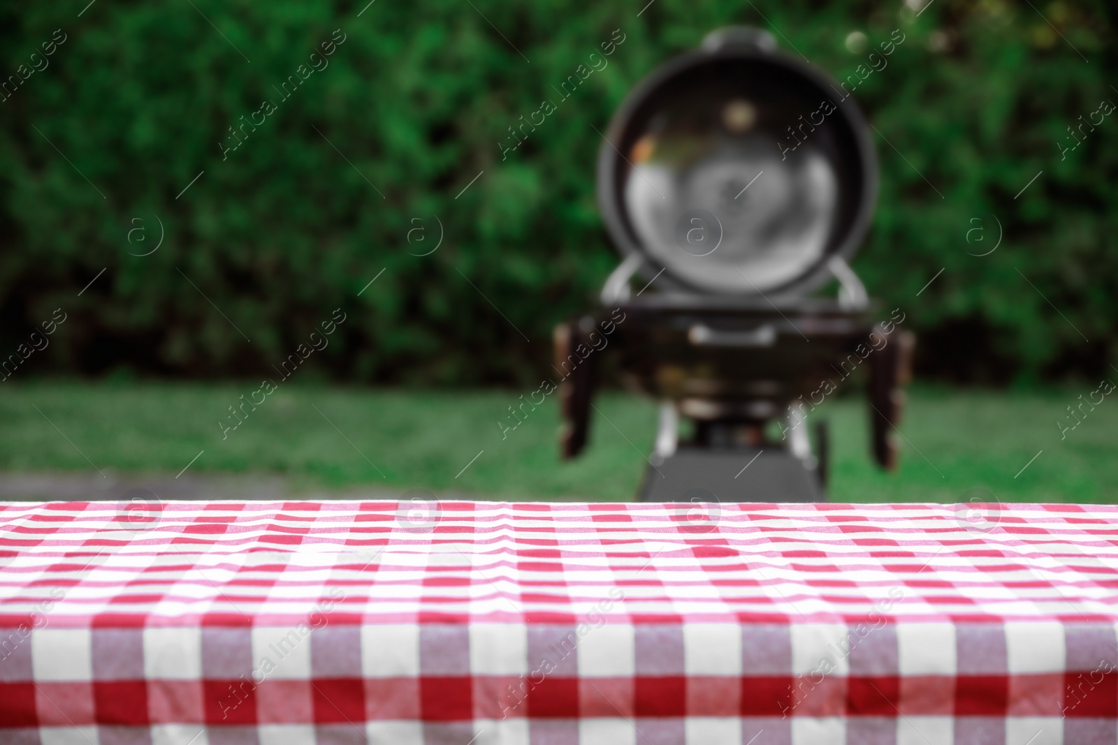 Photo of Picnic table with red checkered cloth and blurred barbecue grill outdoors