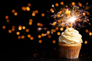 Delicious birthday cupcake with sparkler on black table against blurred lights. Space for text