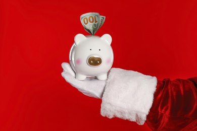 Photo of Santa Claus holding piggy bank with dollar banknotes on red background, closeup