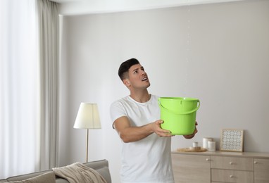 Photo of Emotional man collecting water leaking from ceiling in living room. Damaged roof