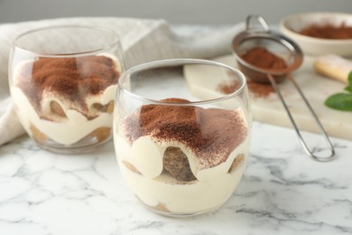 Delicious tiramisu in glasses and sieve with cocoa powder on white marble table, closeup