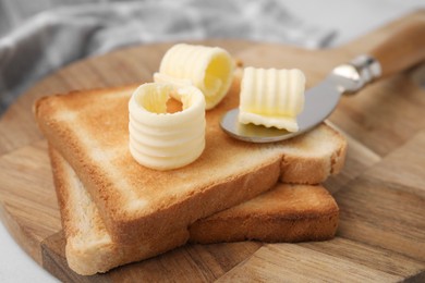 Photo of Tasty butter curls, knife and toasts on wooden board, closeup