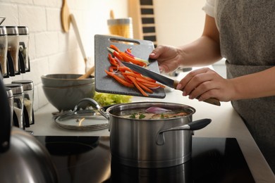 Woman putting cut vegetables into pot with delicious bouillon on stove in kitchen, closeup