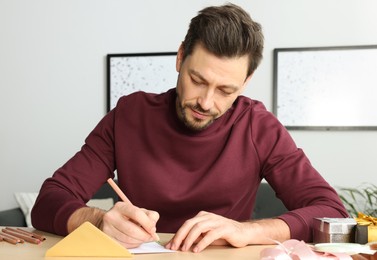 Man writing message in greeting card at wooden table in room