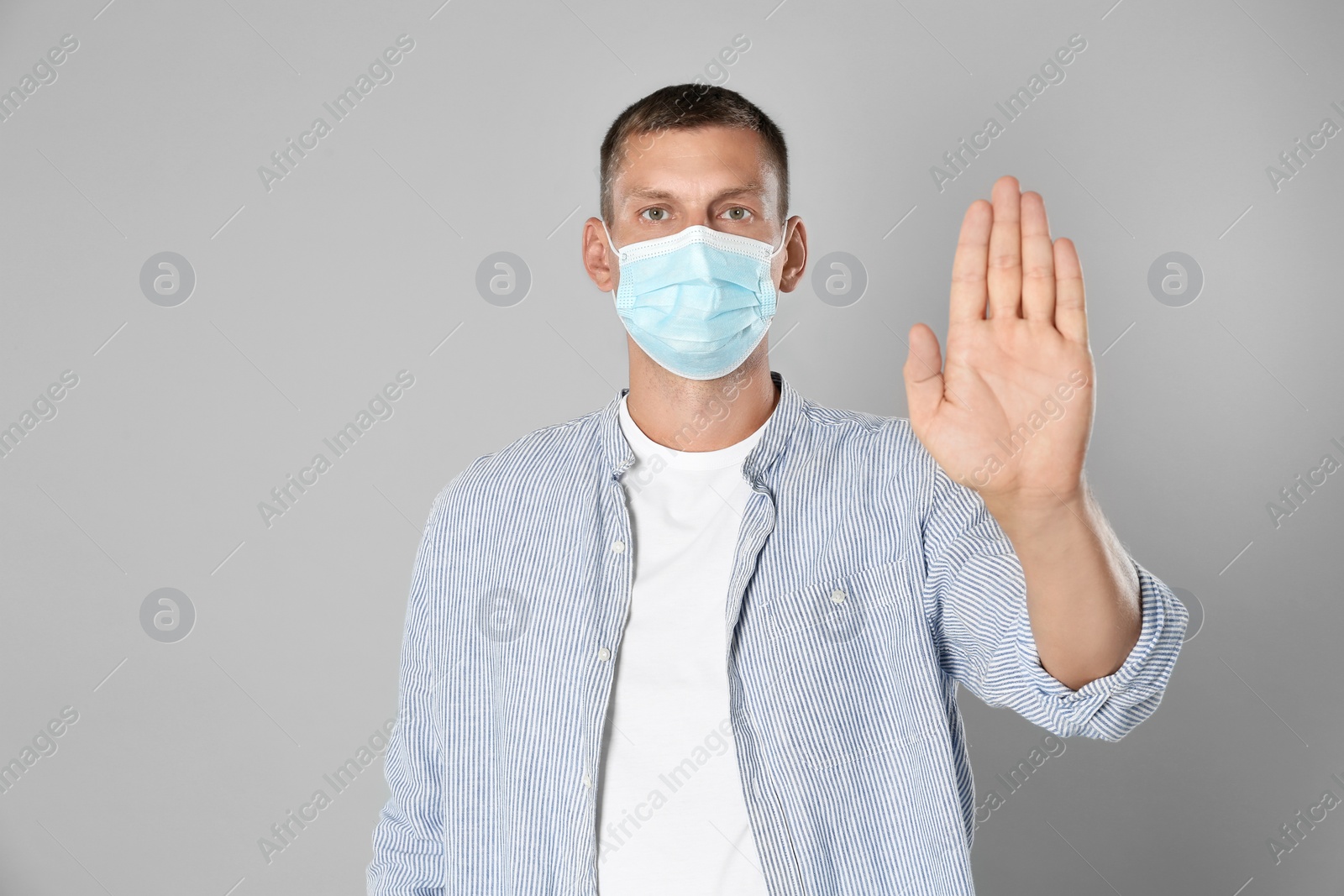 Photo of Man in protective mask showing stop gesture on grey background. Prevent spreading of coronavirus