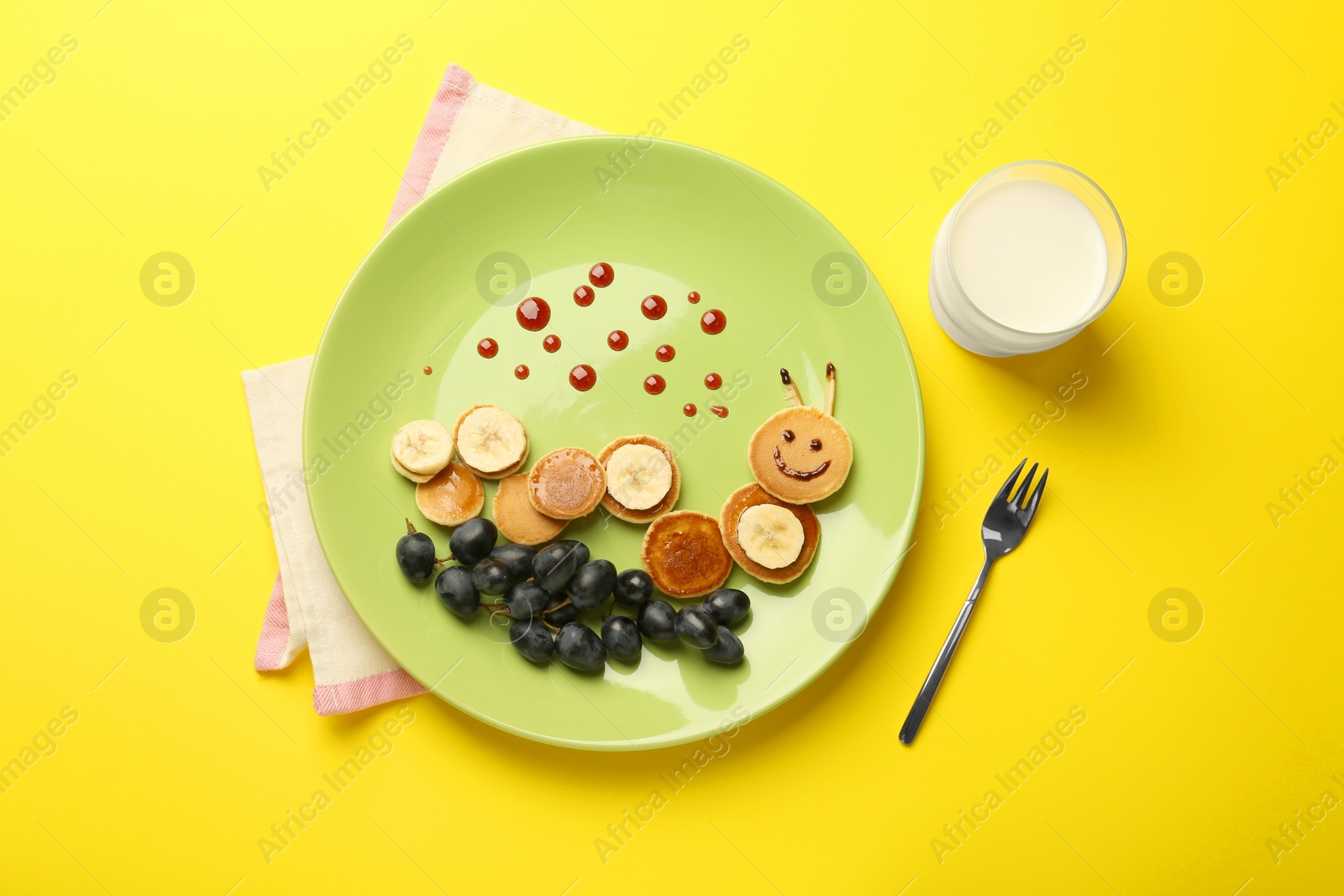 Photo of Creative serving for kids. Plate with cute caterpillar made of pancakes, grapes and banana on yellow background, flat lay