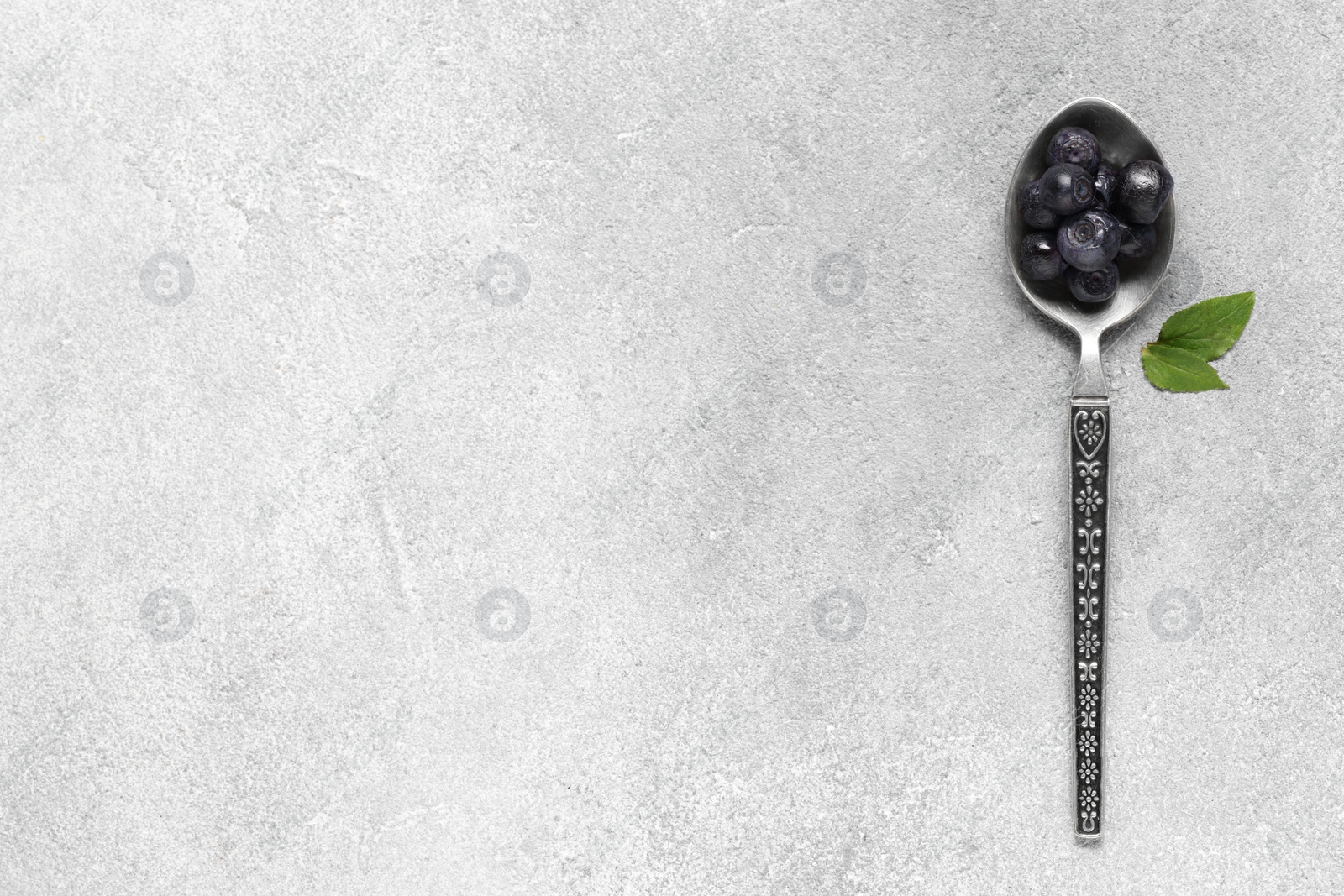 Photo of Spoon with ripe bilberries and leaves on light grey textured table, top view. Space for text