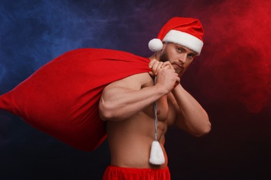 Muscular young man in Santa hat holding bag with presents on color background