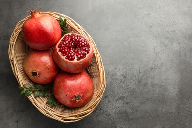 Fresh pomegranates and green leaves in wicker basket on grey table, top view. Space for text