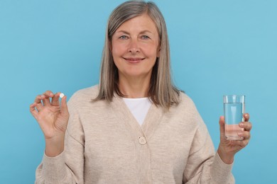 Senior woman with glass of water and pill on light blue background