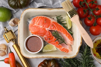 Fresh fish, lime, rosemary and marinade in baking dish surrounded by products on light table, flat lay