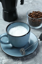 Mini mixer (milk frother), whipped milk in cup and coffee beans on grey textured table