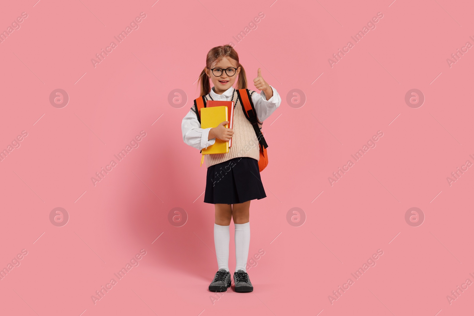 Photo of Happy schoolgirl in glasses with backpack and books showing thumb up gesture on pink background