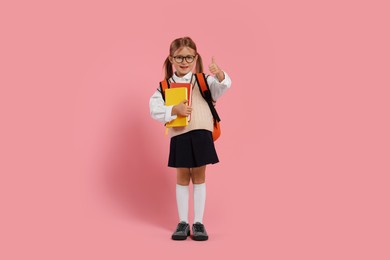 Photo of Happy schoolgirl in glasses with backpack and books showing thumb up gesture on pink background