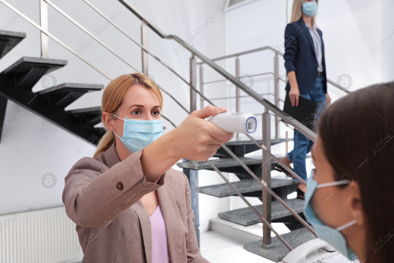 Photo of Woman in mask measuring temperature of employee with noncontact thermometer at office reception