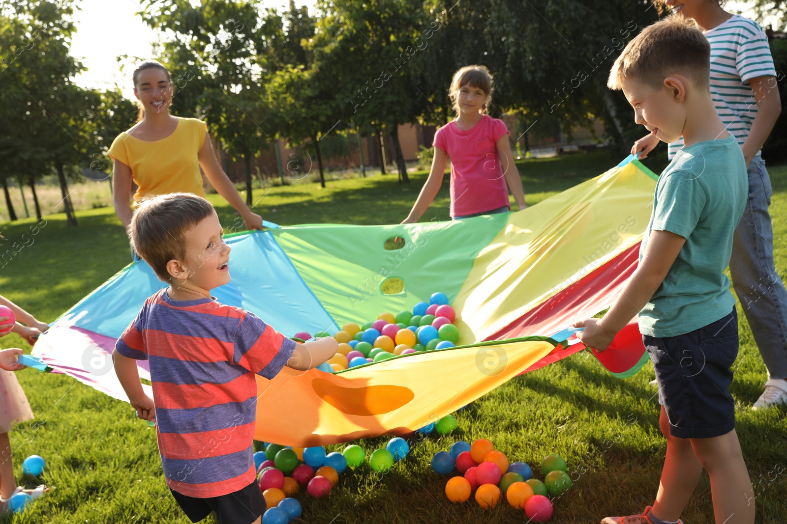 Photo of Group of children and teachers playing with rainbow playground parachute on green grass. Summer camp activity