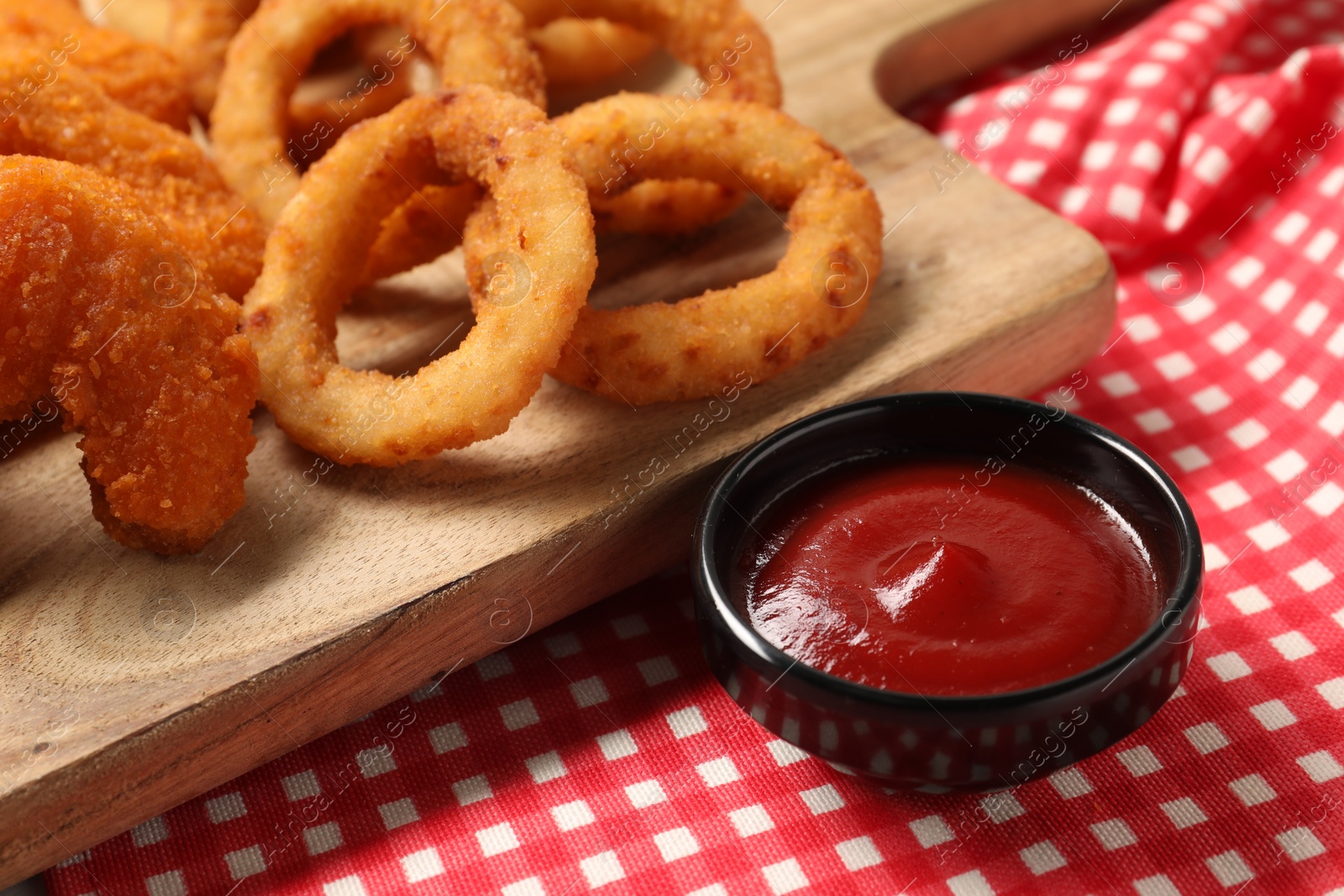 Photo of Tasty ketchup with chicken nuggets and onion rings on checkered cloth, closeup