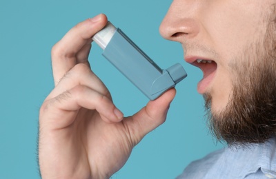 Young man using asthma inhaler on color background, closeup