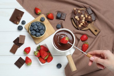 Woman dipping fresh strawberry in fondue pot with melted chocolate at white wooden table, top view