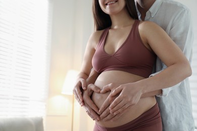 Photo of Pregnant young woman making heart with hands on belly and husband near her at home, closeup. Space for text