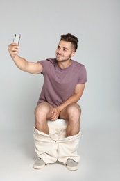 Photo of Young man taking selfie while sitting on toilet bowl against gray background