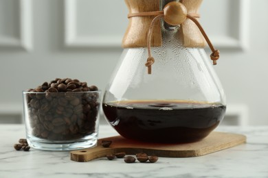 Photo of Glass chemex coffeemaker with coffee and beans in bowl on white marble table, closeup